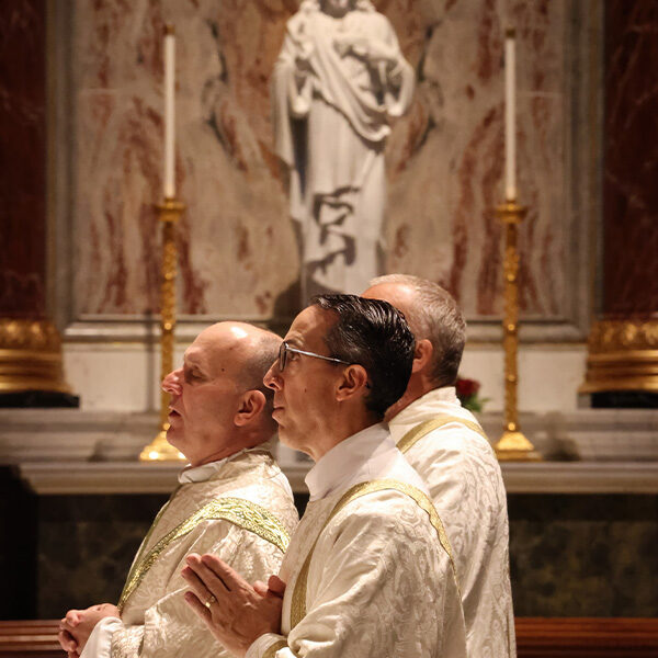 Two Deacons and a priest praying before the altar with the sacred heart of Jesus statue in the background.