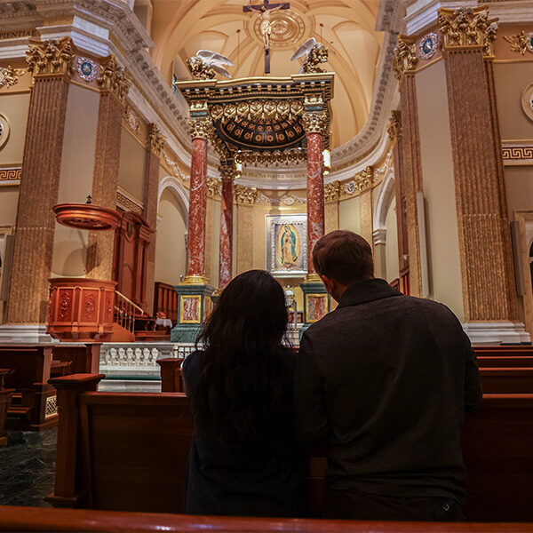 A newlywed couple is kneeling in prayer in the aisle of a church pew at the Shrine.