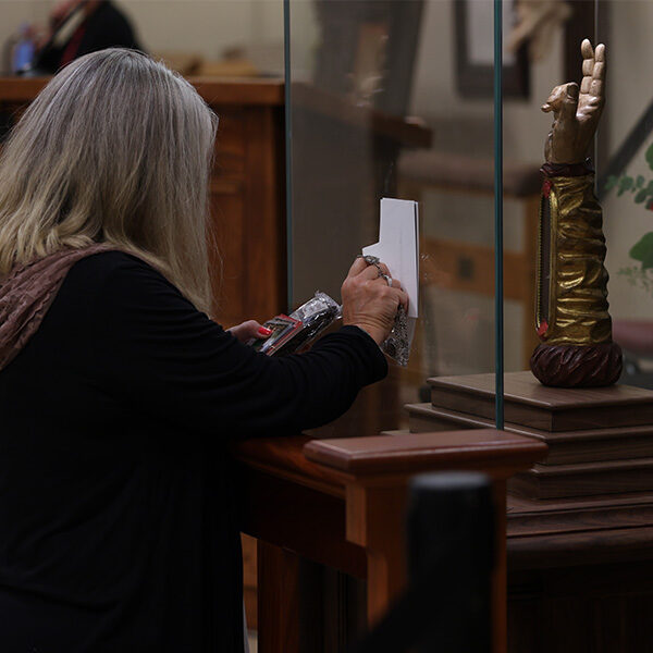 An older woman kneeling to venerate a relic while holding items up to the relic arm of Saint Jude.