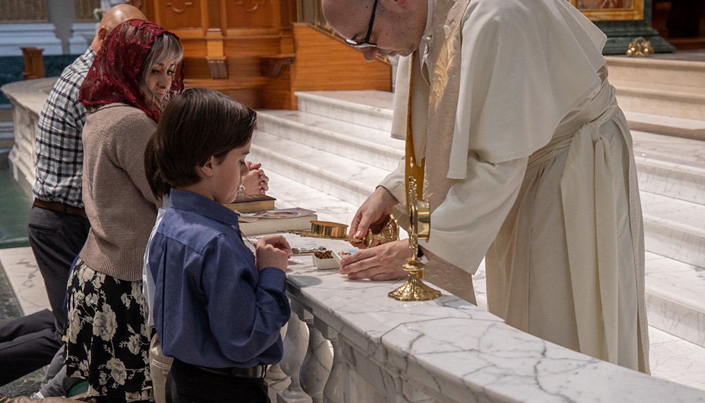 A pilgrimage family of 3 kneeling at the altar in front of a priest at the Shrine in La Crosse, Wisconsin.