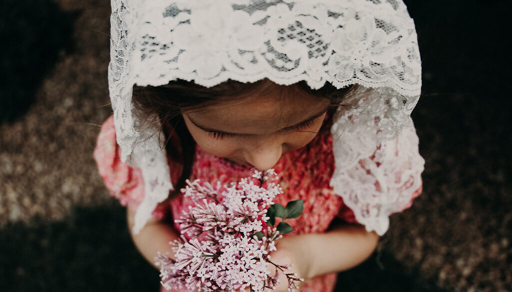 A young girl wearing a white floral veil while holding pink dainty flowers.