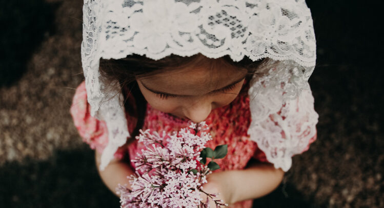 A young girl wearing a white floral veil while holding pink dainty flowers.