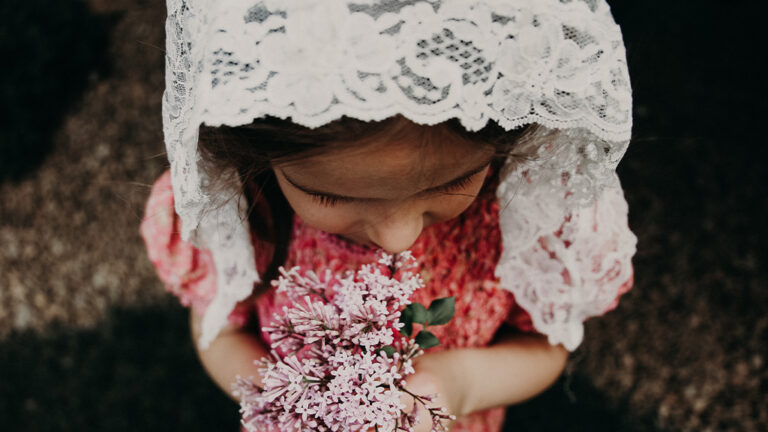 A young girl wearing a white floral veil while holding pink dainty flowers.