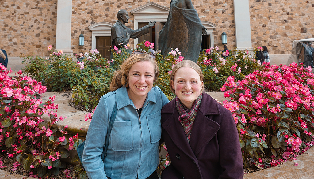 Sarah Greydanus and a companion visiting the Shrine of Our Lady of Guadalupe during a fall pilgrimage, surrounded by vibrant pink flowers.