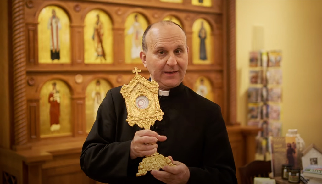 Father Paul Check holding a golden reliquary in a room adorned with sacred artwork, featured in a video titled 'Blessed Carlo Acutis: Living the Christian Life' at the Shrine of Our Lady of Guadalupe in La Crosse, Wisconsin.