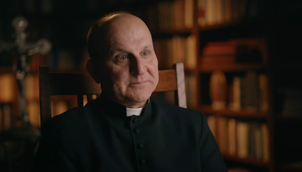 Father Paul Check seated in a warmly lit library, speaking as part of a video titled 'Love, Joy, and the Commandments' at the Shrine of Our Lady of Guadalupe in La Crosse, Wisconsin.