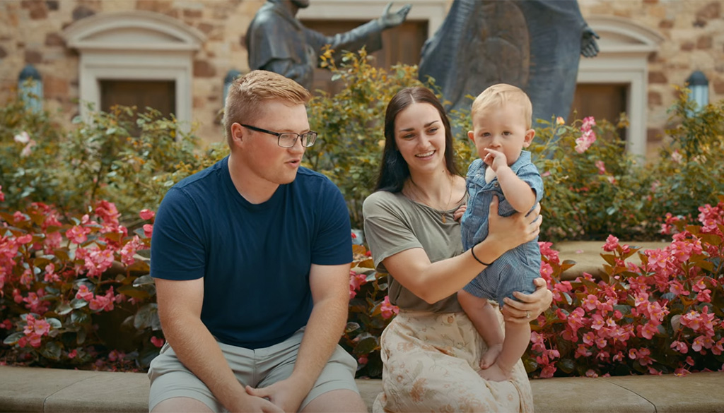 A family sitting in a garden with vibrant flowers at the Shrine of Our Lady of Guadalupe in La Crosse, Wisconsin, featured in a video titled 'Finding the Answer to Prayer'.